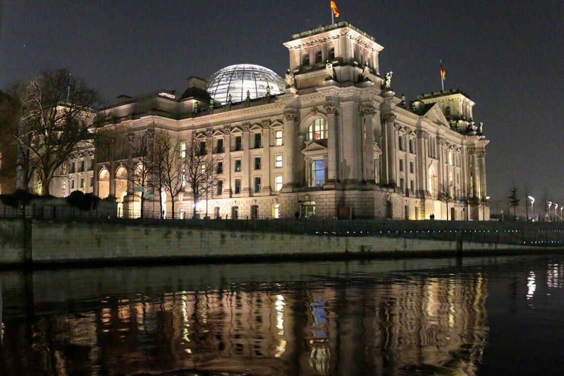 Der angestrahlte Rechstag in Berlin spiegelt sich bei Nacht im Wasser.